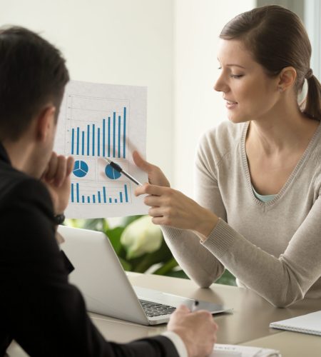 Attractive millennial businesswoman presenting company financial indicators, telling business partner about investment plan, showing document with project positive stats sitting at office desk